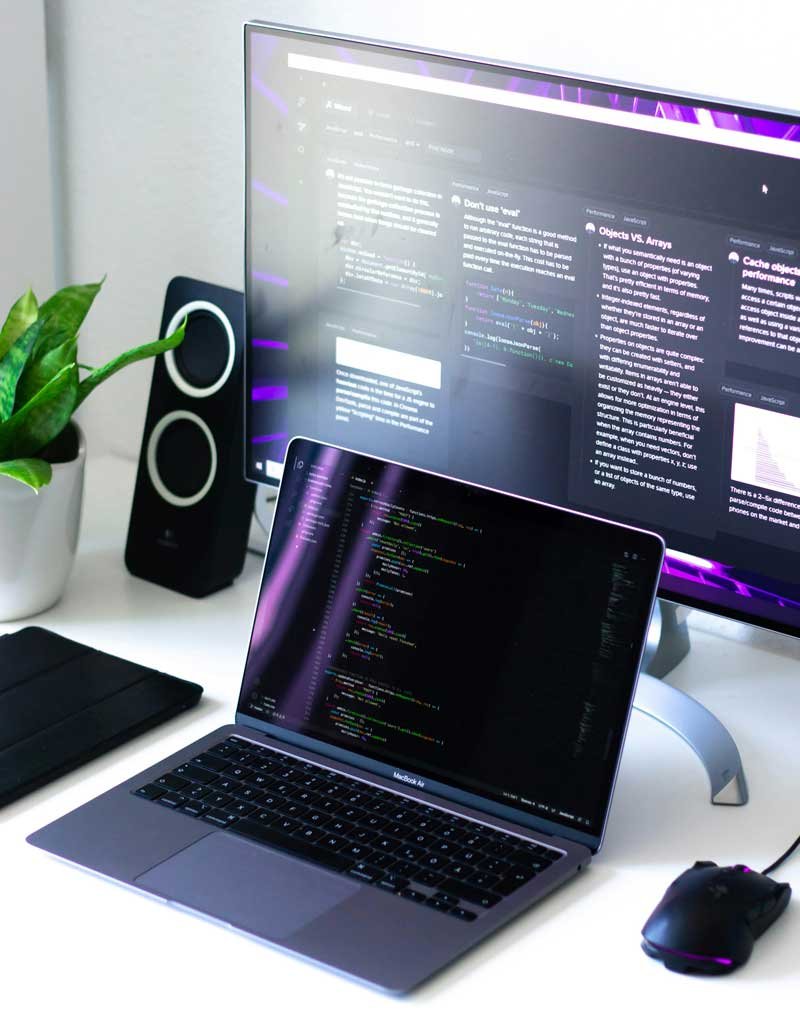 Notebook sob uma mesa branca, com mouse gamer ao lado e monitor auxiliar ao fundo. Ambas as telas possuem códigos em linguagem de programação.